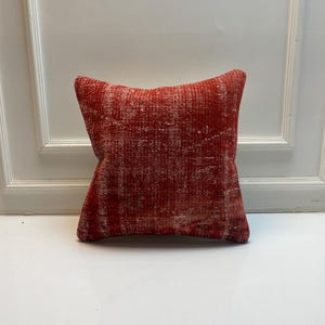 May include: A square throw pillow featuring a distressed, weathered texture in shades of deep red and white. The fabric displays a faded, worn appearance with visible variations in color density, creating a mottled, vintage-inspired surface pattern.