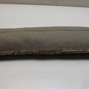 May include: A long, rectangular pillow with a muted olive-green top and a patterned bottom. The top fabric is smooth, while the bottom features a textured, multi-colored design. The pillow is designed for decorative use.