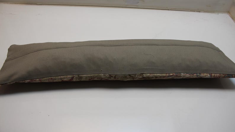 May include: A long, rectangular pillow with a muted olive-green top and a patterned bottom. The top fabric is smooth, while the bottom features a textured, multi-colored design. The pillow is designed for decorative use.