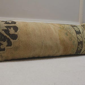 May include: Rectangular beige pillow with a woven texture and dark brown and teal geometric patterns on the ends. The pillow is leaning against a white wall. Decorative home decor.