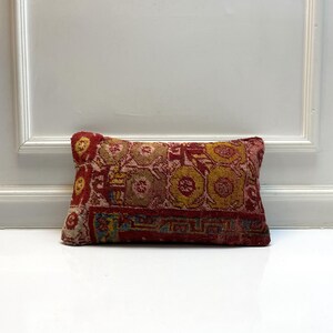 May include: A rectangular pillow with a detailed, colorful pattern. The pillow features a red background with yellow, blue, and beige accents. The design includes floral and geometric shapes, creating a vintage or ethnic aesthetic.