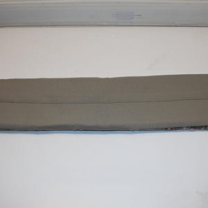 May include: A rectangular, light gray fabric item with a seam down the middle. The bottom edge features a patterned design. The item is likely a car accessory, possibly a sun visor or a similar component.