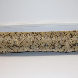 May include: A long, rectangular pillow with a beige and brown patterned design. The pillow has a distressed, vintage look, with a repeating geometric pattern in darker shades. The pillow is placed on a white surface.