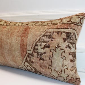 May include: A rectangular lumbar pillow featuring a woven textile cover with a faded, distressed appearance. The fabric displays a muted color palette of beige, tan, and soft terracotta, accented by a dark brown geometric pattern on one side.
