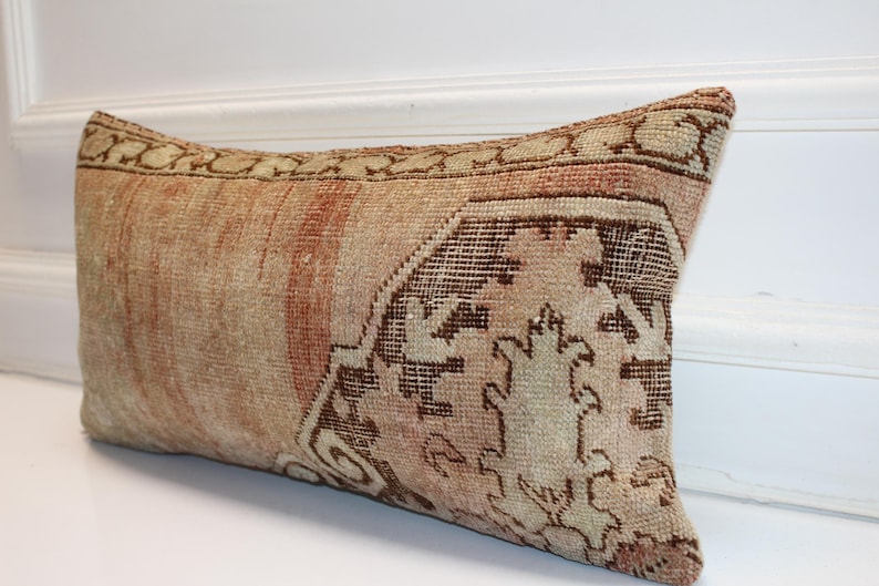 May include: A rectangular lumbar pillow featuring a woven textile cover with a faded, distressed appearance. The fabric displays a muted color palette of beige, tan, and soft terracotta, accented by a dark brown geometric pattern on one side.