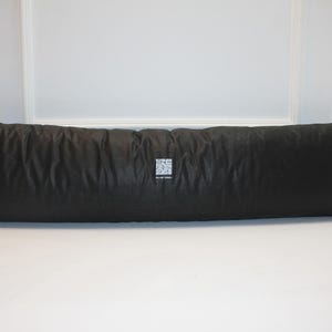 May include: A long, rectangular black pillow with a textured surface. The pillow is positioned horizontally and appears to be made of a soft, pliable material. A small, square white tag with a QR code is visible on the pillow's surface.