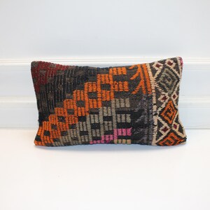 Puede incluir: Cojín rectangular decorativo con un patrón geométrico en tonos naranja, negro, gris y rosa. El cojín tiene un aspecto texturizado y está colocado sobre un fondo blanco.