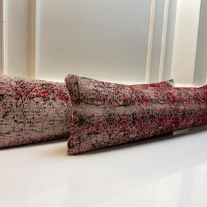 Op de afbeelding: Twee rechthoekige kussens met een versleten, vintage uitstraling. De kussens hebben een rood en zwart patroon op een crèmekleurige achtergrond. Het patroon heeft een traditioneel, sierlijk ontwerp. De kussens liggen op een wit oppervlak.