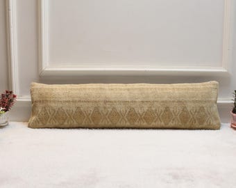 Peut inclure: Un long coussin rectangulaire beige avec un motif géométrique en losanges. Le coussin est posé sur une surface blanche, avec des plantes décoratives en arrière-plan. Les dimensions du coussin ne sont pas visibles.