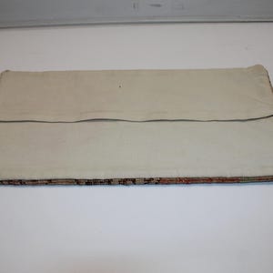 May include: A rectangular pillow cover featuring a plain off-white canvas back with a horizontal fabric flap closure. The bottom edge reveals a glimpse of a multi-colored woven textile pattern with intricate geometric details in brown and orange.