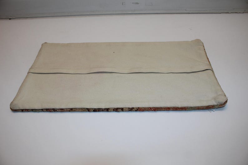 May include: A rectangular pillow cover featuring a plain off-white canvas back with a horizontal fabric flap closure. The bottom edge reveals a glimpse of a multi-colored woven textile pattern with intricate geometric details in brown and orange.
