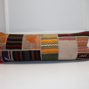 May include: A rectangular lumbar pillow featuring a patchwork design composed of various woven textile segments. The fabric panels display a diverse array of colors including orange, black, yellow, green, beige, and pink, with striped and geometric patterns.