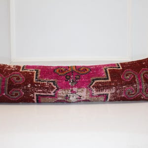 Puede incluir: Un cojín decorativo largo y rectangular con un aspecto vintage y desgastado. El cojín presenta un diseño geométrico central rosa y naranja, enmarcado por un borde burdeos oscuro con volutas rosas. El cojín está colocado sobre una superficie blanca.