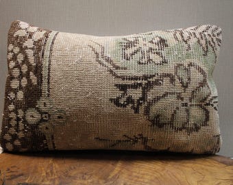 May include: Rectangular decorative pillow with a vintage rug design. The pillow features a beige background with floral patterns in shades of brown and green. The pillow is placed on a wooden surface.