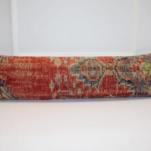 May include: A long, rectangular lumbar cushion featuring a distressed, woven textile cover. The fabric displays a vibrant red base colour with intricate, multi-coloured geometric and floral patterns in shades of blue, beige, and green throughout the surface.