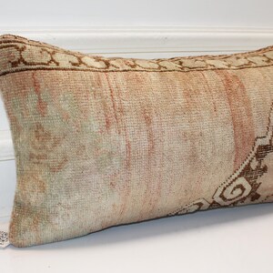 May include: A rectangular lumbar pillow featuring a faded, vintage-style woven textile pattern in muted shades of beige, soft peach, and dark brown. The fabric displays a textured, knotted appearance with a decorative border and intricate motifs.