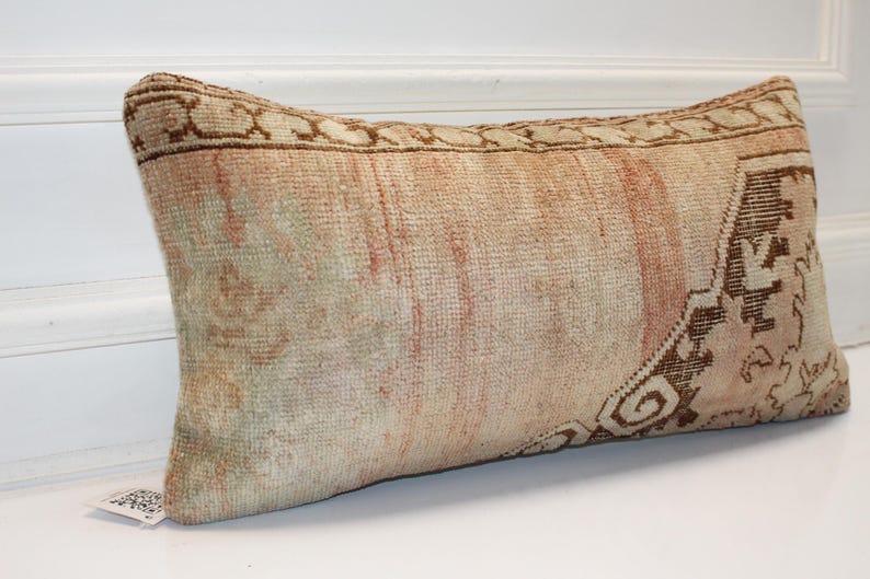 May include: A rectangular lumbar pillow featuring a faded, vintage-style woven textile pattern in muted shades of beige, soft peach, and dark brown. The fabric displays a textured, knotted appearance with a decorative border and intricate motifs.