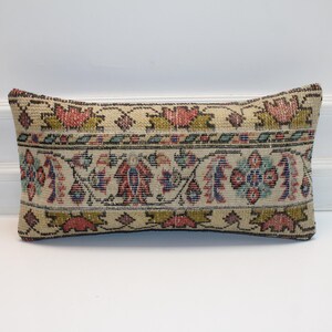 May include: Rectangular beige pillow with a detailed floral pattern in red, blue, and green. The pillow has a vintage, textured appearance, suggesting it may be handmade or antique. The design is symmetrical, with a border.