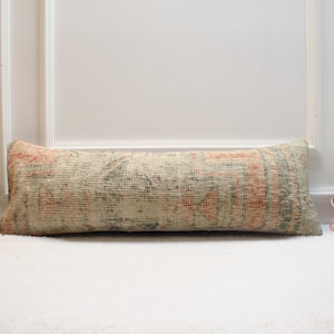 May include: A long, rectangular pillow with a vintage-style patterned fabric. The pillow is beige with shades of pink, green, and brown. It is lying on a white surface.