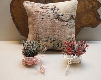 Peut inclure: Un coussin décoratif avec un motif floral délavé dans les tons beige, marron et sarcelle. Deux jardinières miniatures en forme de vélo, une rose et une blanche, contiennent de petites plantes à fleurs rouges et vertes. En arrière-plan, une tranche de bois.