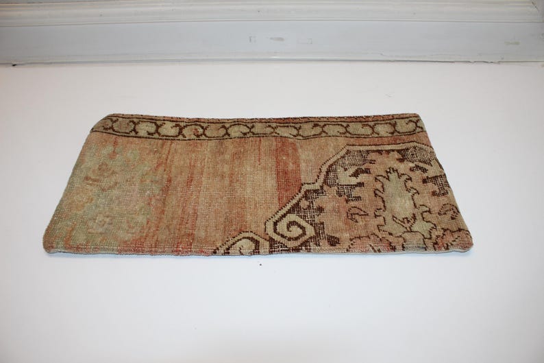 May include: A rectangular pillow cover featuring a faded, woven textile pattern in shades of muted orange, beige, and dark brown. The design includes a decorative border with a repeating scroll motif and a central area with intricate floral elements.