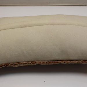 May include: A rectangular throw pillow featuring a solid off-white fabric on the top side with a visible horizontal seam. The bottom edge displays a textured, woven border in a warm, earthy brown tone with a repeating geometric pattern.