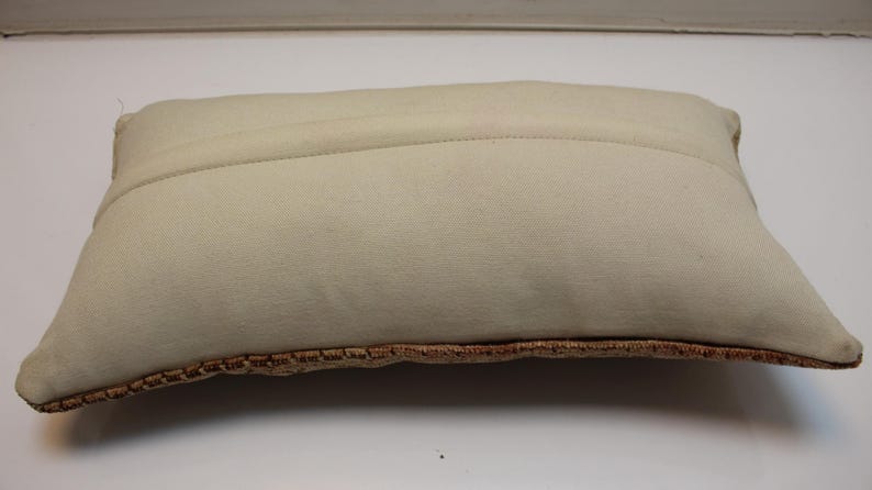 May include: A rectangular throw pillow featuring a solid off-white fabric on the top side with a visible horizontal seam. The bottom edge displays a textured, woven border in a warm, earthy brown tone with a repeating geometric pattern.