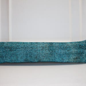 May include: A long, rectangular turquoise pillow with a distressed, textured appearance. The pillow is placed on a white surface, with a white wall in the background. The pillow's dimensions are not visible.