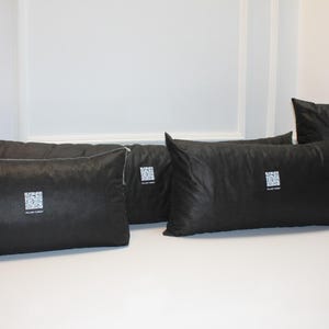 May include: Several black pillows of varying sizes are arranged on a white surface. Each pillow has a square, white graphic and text. The pillows are rectangular and square, with a smooth, dark fabric.
