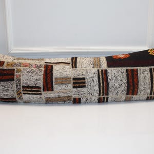 May include: Long, rectangular cushion with a patchwork design. The cushion features a mix of neutral colours, including beige, brown, and black, with geometric patterns. The cushion is approximately 100 cm long.