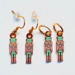 Può includere: Set di tre orecchini a forma di schiaccianoci dorati. Due orecchini sono di tipo pendente con un ciondolo a forma di schiaccianoci verde e rosa. Il terzo orecchino è un cerchio con un ciondolo a forma di schiaccianoci verde e rosa.