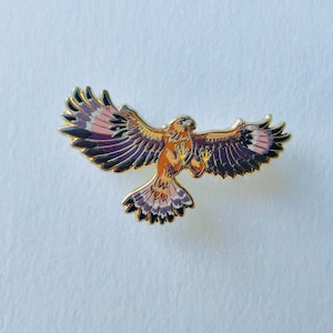 May include: A gold-toned metal enamel pin shaped like a hawk in flight with wings spread wide. The bird features a color palette of orange, deep purple, and soft pink, with gold outlines defining the feathers and body structure.