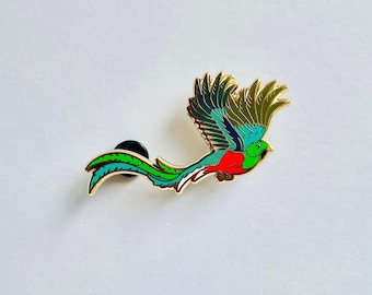 May include: A gold-toned metal enamel pin shaped like a quetzal bird in flight. The bird features vibrant green, teal, and red plumage with detailed gold outlines and a long, flowing tail. A black circular pin backing is visible on the reverse side.