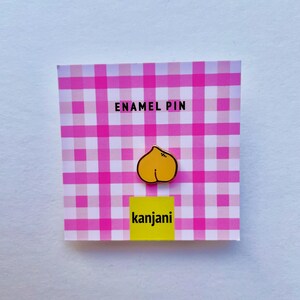Può includere: Spilla smaltata con un design a pesca gialla con contorno nero, su sfondo a quadri rosa e bianchi. Le parole "ENAMEL PIN" sono in alto e "kanjani" in basso su un rettangolo giallo.