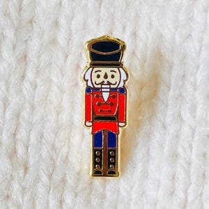 May include: A metal enamel pin shaped like a nutcracker soldier. The figure wears a black hat, a red jacket with gold-coloured trim, and blue trousers. The design features bold black outlines and a white face with a painted moustache and beard.