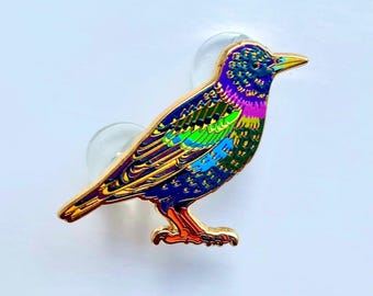 Puede incluir: Un broche de esmalte metálico con forma de pájaro, con un acabado iridiscente vibrante en tonos morado, azul, verde y amarillo. El pájaro tiene un contorno de metal dorado y dos cierres de goma transparentes detrás del cuerpo.