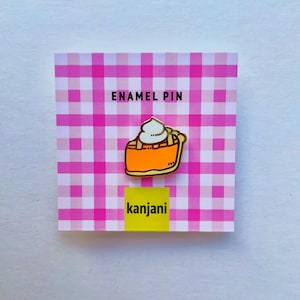 Könnte beinhalten: Ein Emaille-Pin mit einem Stück Kürbiskuchen mit Schlagsahne, vor einem rosa-weiß karierten Hintergrund. Der Pin ist orange, gelb und weiß mit dem Text "ENAMEL PIN" und "kanjani".