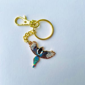 Op de afbeelding: Een goudkleurige sleutelhanger met een ekster met uitgespreide vleugels. De vogel heeft een zwart lichaam, witte vleugelaccenten en iriserende blauwe en groene staartveren. De sleutelhanger bevat een sluiting en een ring.