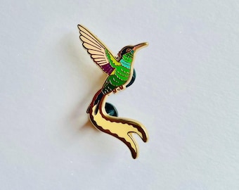 May include: A gold-toned metal enamel pin shaped like a hummingbird in flight. The bird features vibrant green, purple, and light yellow enamel detailing on its wings and body, with a long, curved tail and a pointed beak in a gold finish.