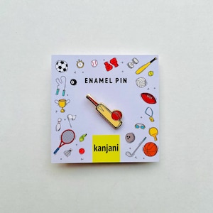 May include: An enamel pin featuring a cricket bat and ball, with a yellow and red design. The pin is on a white card with sports-themed illustrations and the text "ENAMEL PIN" and "kanjani".