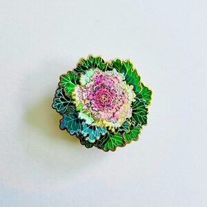 以下が含まれることがあります： 花キャベツの形をした装飾的なエナメルピン。ピンは、緑、ピンク、金色の詳細なデザインが特徴です。葉は金の縁取りのある緑色で、中央はピンクと白です。