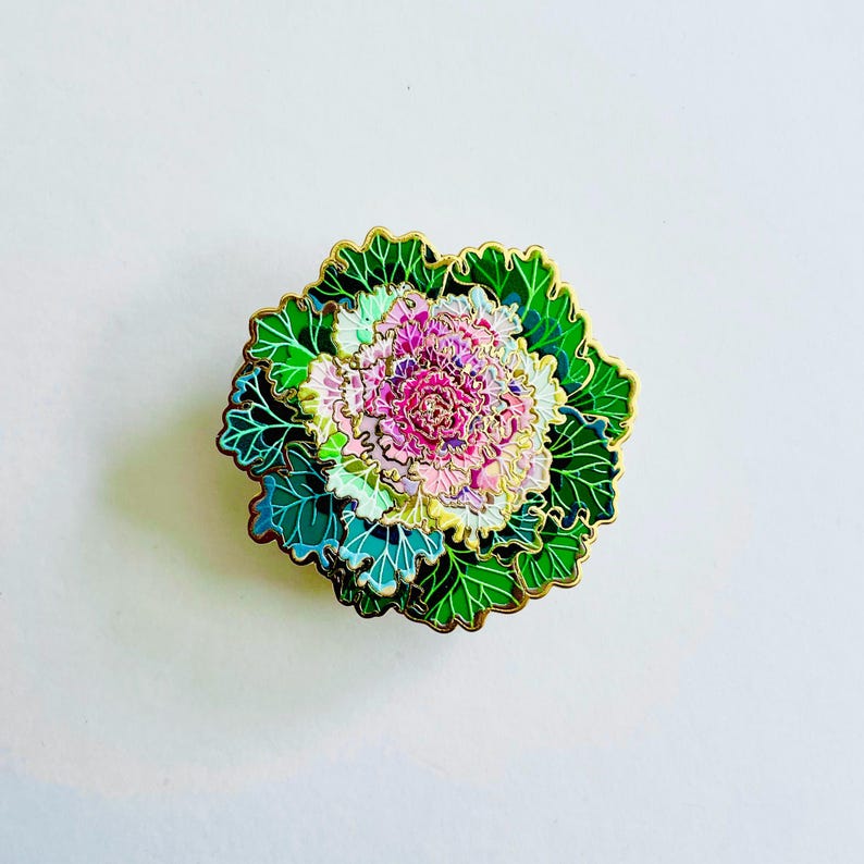 以下が含まれることがあります： 花キャベツの形をした装飾的なエナメルピン。ピンは、緑、ピンク、金色の詳細なデザインが特徴です。葉は金の縁取りのある緑色で、中央はピンクと白です。