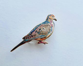 Puede incluir: Un broche de esmalte en tono dorado con forma de paloma huilota. El ave presenta detalles intrincados de plumas en tonos azul, rosa, marrón y crema, con un contorno de metal dorado pulido que define las alas, el cuerpo, la cabeza y la cola larga.