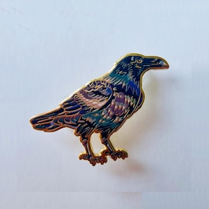 May include: An enamel pin featuring a raven design. The bird is rendered in shades of dark blue and purple, with gold-coloured outlines. The pin showcases a detailed representation of a raven, with intricate feather detailing.
