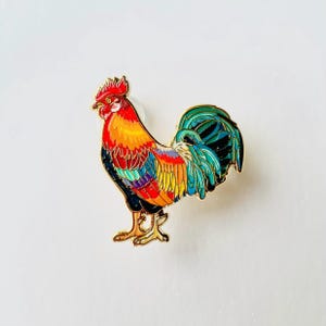 May include: A colourful rooster pin with a gold outline. The rooster has a red comb, yellow and orange body, and a vibrant blue and green tail. The pin is set against a white background.