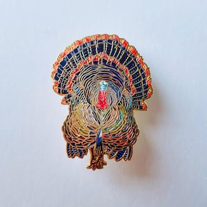 Può includere: Una spilla a forma di tacchino colorato con contorno dorato. La spilla presenta un design dettagliato con piume blu, rosse e dorate. La testa del tacchino è rossa e blu, e il corpo ha un mix di colori.