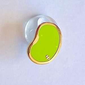 Peut inclure: Une broche en émail en forme de haricot avec un centre vert lime vif entouré d'un bord en métal doré poli. La surface de la broche est lisse et brillante, avec un petit détail distinctif à trois points gravé dans le coin inférieur droit.