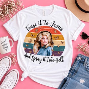 Puede incluir: Una camiseta blanca con un gráfico de una persona rubia con camisa de mezclilla centrada en un círculo de atardecer retro con mariposas. El texto dice "Tease it to Jesus" arriba y "And Spray it Like Hell" debajo del gráfico.