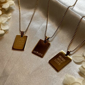 Könnte beinhalten: Drei goldene Halsketten mit rechteckigen Anhängern. Die Anhänger tragen folgende Aufschriften: "Love Each Other More", "Always" und "More Self Love".
