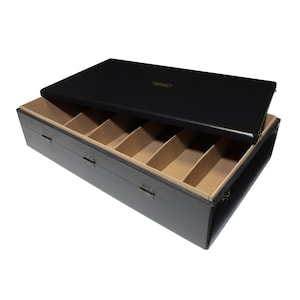 Op de afbeelding: Een zwarte houten kist met een scharnierend deksel, gedeeltelijk open om interne compartimenten te onthullen. De binnenkant is bekleed met een beige stof. Het deksel heeft een klein goudkleurig rechthoekig label. De kist is rechthoekig en heeft metalen sluitingen.